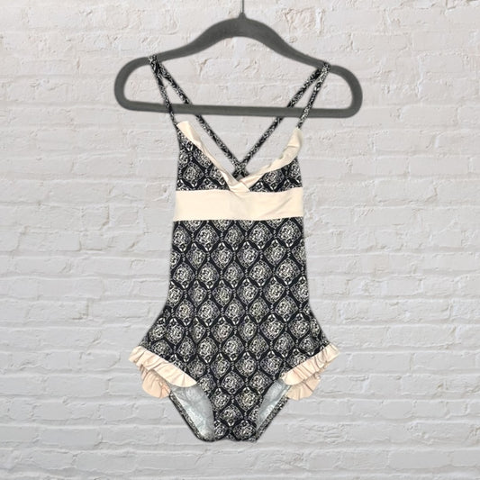 Women's one-piece swimsuit with a black and cream pattern, featuring ruffles and cross-back straps, hanging on a black hanger.