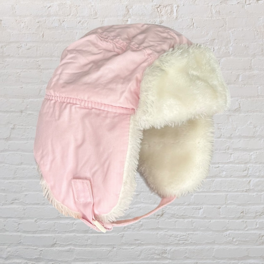 Pink trapper hat with white faux fur lining and ear flaps against a white brick background.