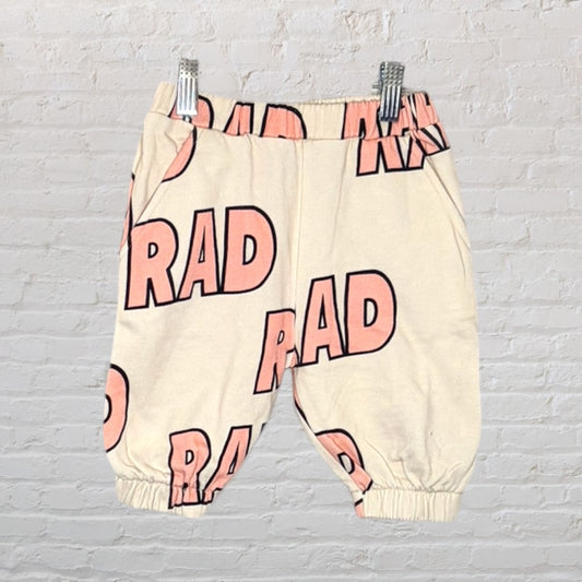 Cream-colored pants with pink 'RAD' text, featuring an elastic waist and cuffs, displayed against a white brick background.