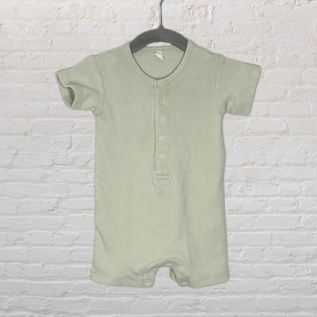 Light green short sleeve henley baby romper with button placket, hanging on a dark hanger against a white brick wall.
