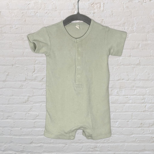 Light green short sleeve henley baby romper with button placket, hanging on a dark hanger against a white brick wall.