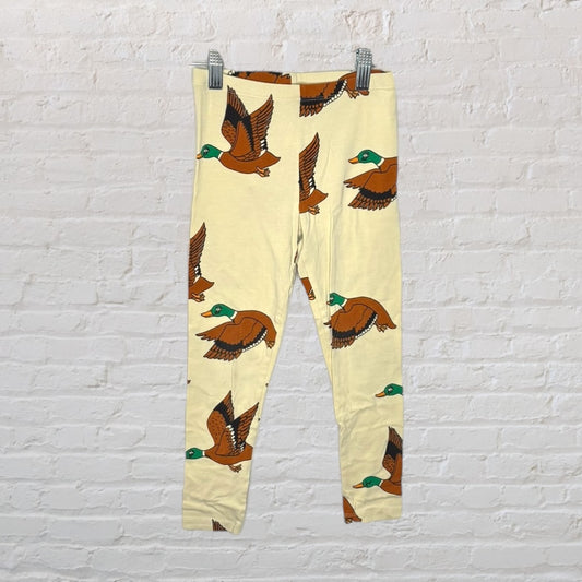 Cream colored leggings with brown and green duck print, hanging on a clip hanger against a brick wall.