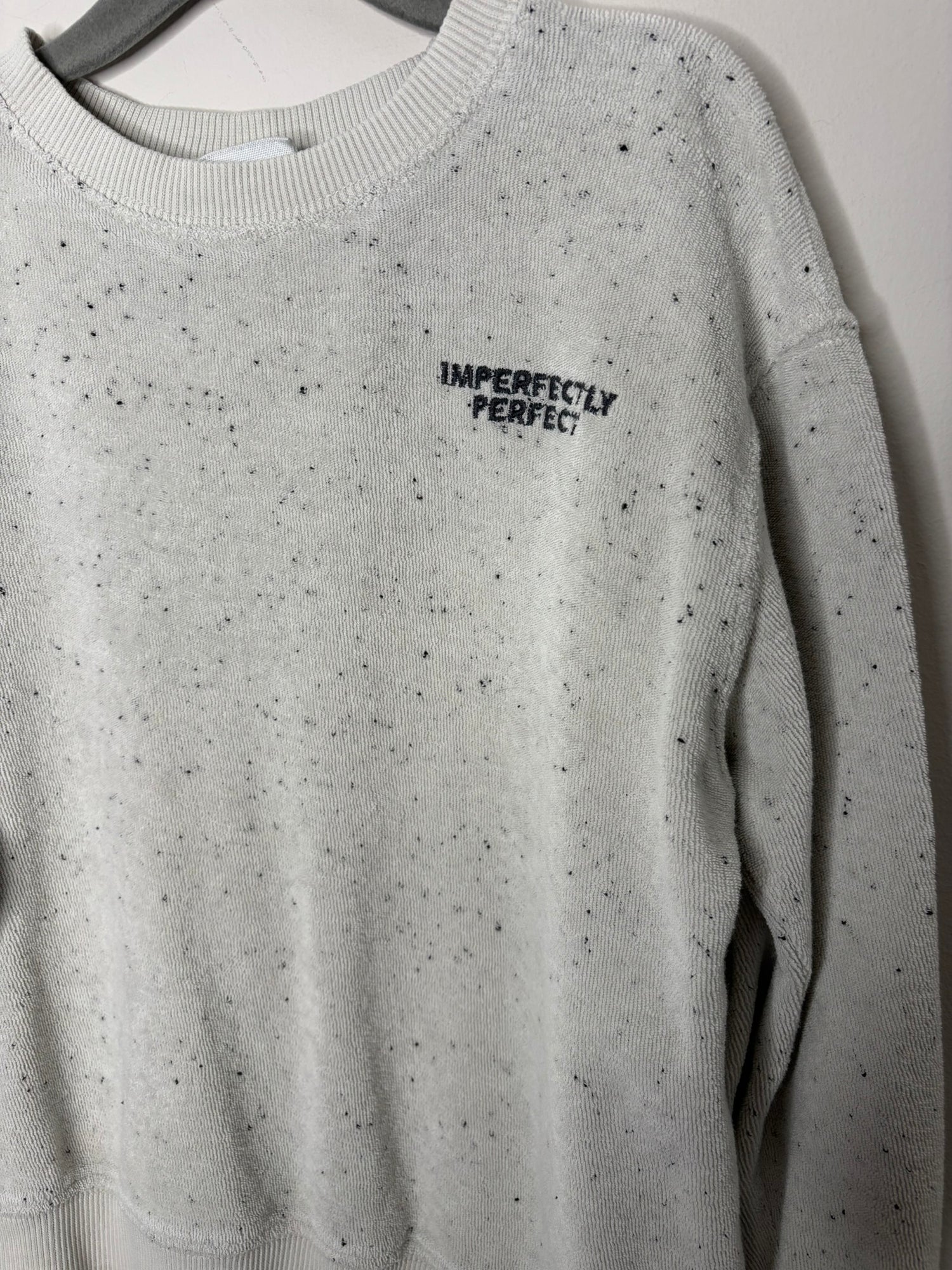 Light grey speckled sweatshirt with 'Imperfectly Perfect' text on the chest, displayed on a hanger against a brick wall.