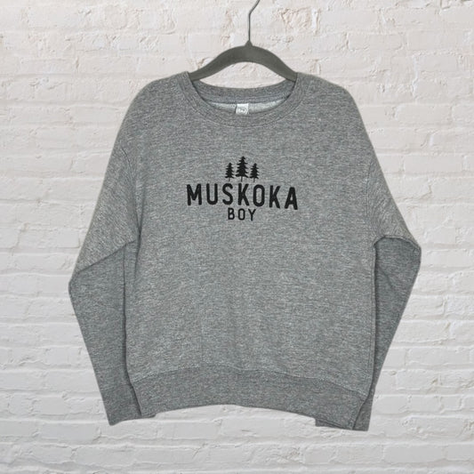 Grey sweatshirt with 'Muskoka Boy' printed in black text, hanging on a hanger against a white brick wall.