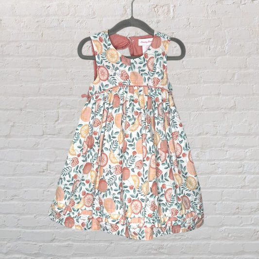 A sleeveless dress with a colorful fruit and leaf print, featuring a fitted bodice and flared skirt, hanging on a hanger against a brick wall.