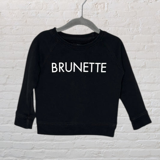 Black sweatshirt with 'Brunette' text in white, displayed on a hanger against a white brick wall.
