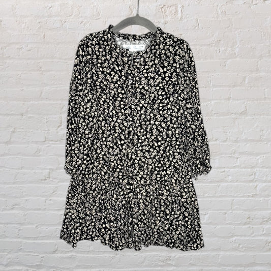 Black dress with white floral pattern and buttons down the front on a hanger against a brick wall.