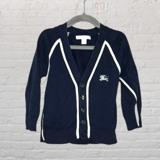 Navy cardigan with white contrast trim and buttons, featuring a subtle logo on the chest, hanging against a white brick background.