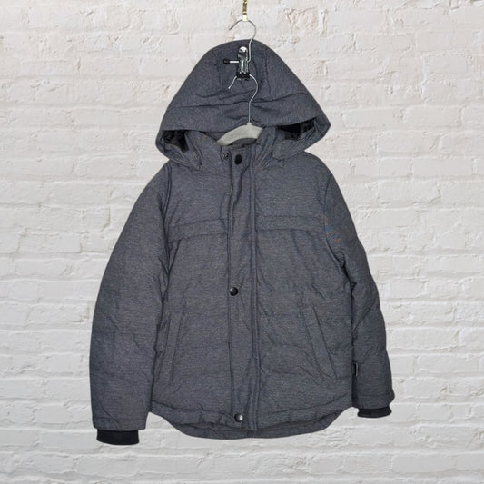 Charcoal grey hooded puffer jacket with button and zipper front, displayed against a white brick wall.