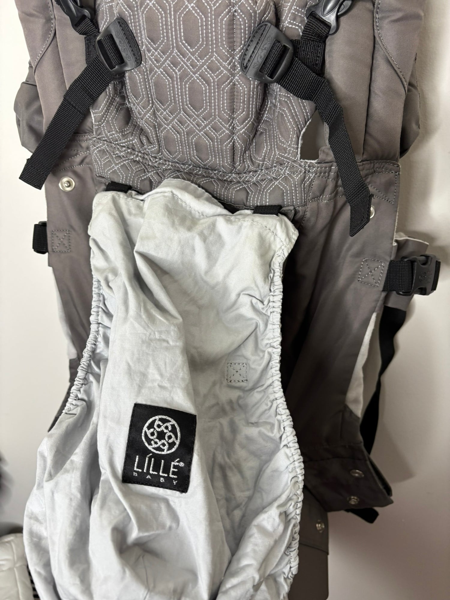Gray ergonomic baby carrier with multiple straps and support, hanging on a hanger.