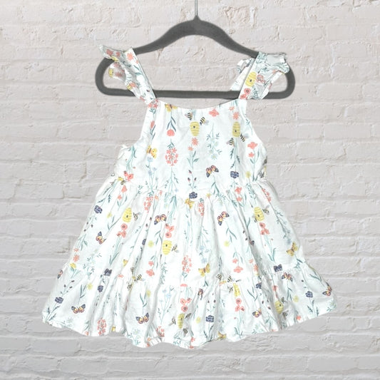 White dress with multicolored floral print and ruffle sleeves hanging on a dark hanger against a brick wall.