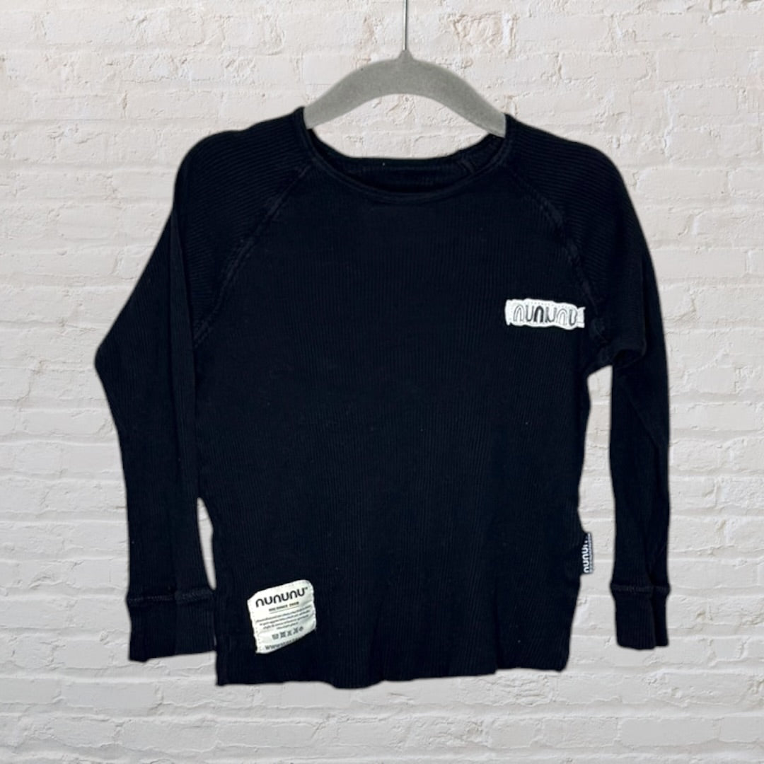 Black long sleeve shirt with Nununu branding on the chest and hem, hung on a grey hanger against a white brick wall.