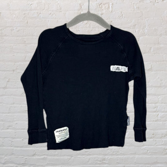 Black long sleeve shirt with Nununu branding on the chest and hem, hung on a grey hanger against a white brick wall.