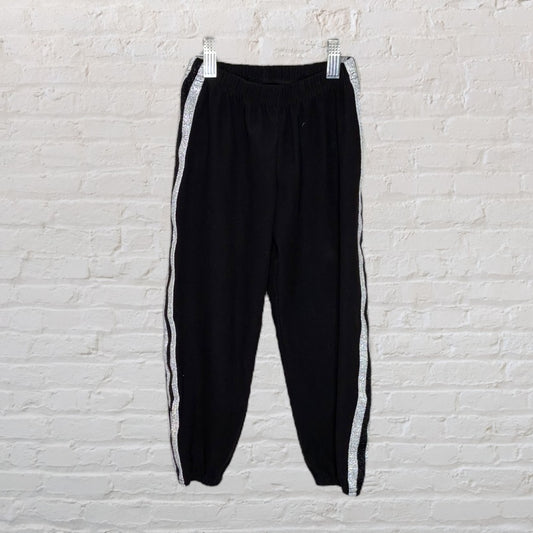 Black jogger pants with elastic waistband and sparkly silver side stripes, hanging against a white brick wall.