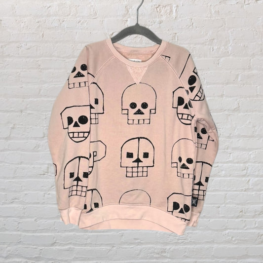 Light pink sweatshirt with black skull prints, displayed against a white brick wall.