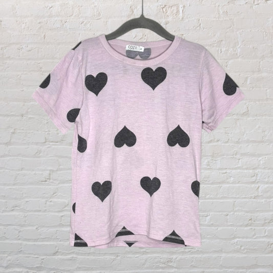 Light pink t-shirt with black heart patterns by Cozii, hanging on a hanger against a brick wall.