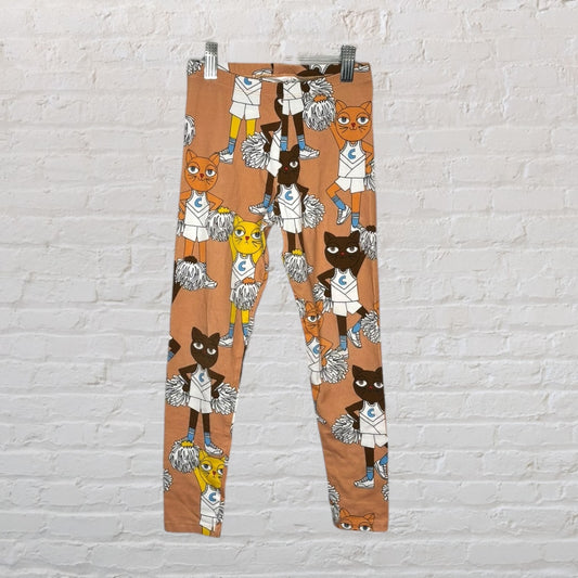 Brown leggings with a colorful cheerleader cats print, hanging against a white brick wall.