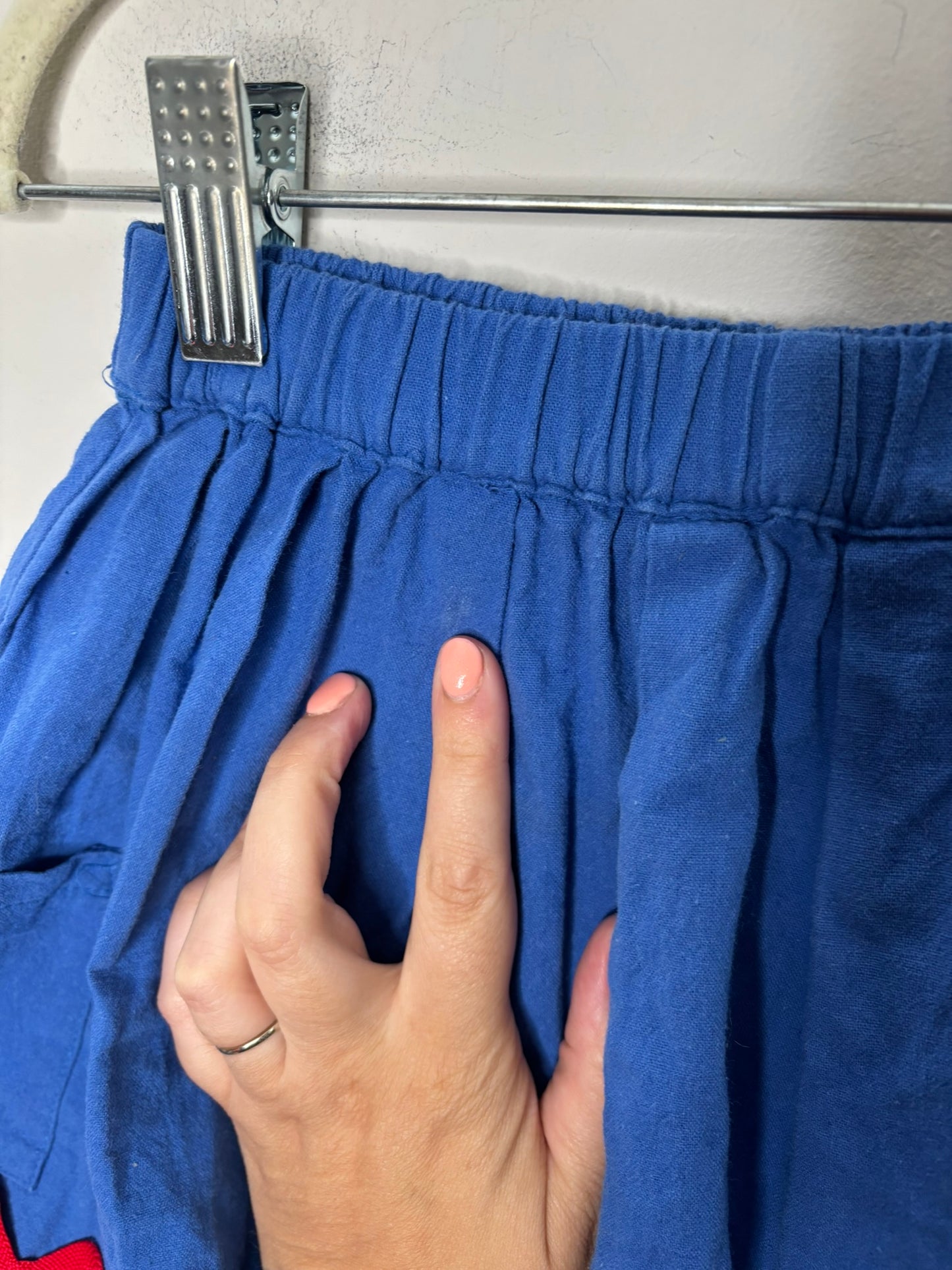 Blue skirt with sun embroidery and red wavy trim, featuring side pockets, displayed on a hanger against a white brick wall.