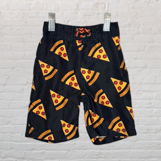 Black swim trunks with a playful pizza slice pattern and a red drawstring, hanging against a white brick wall.