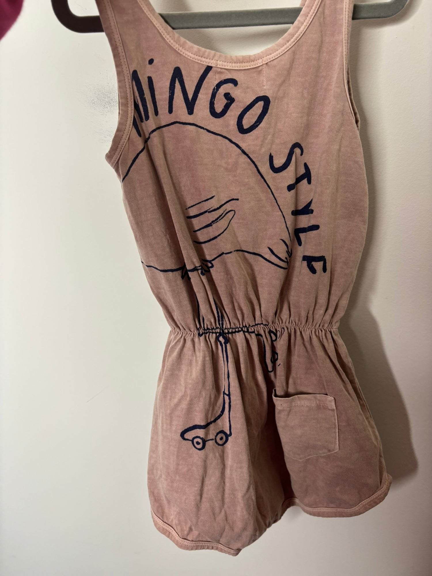 Beige sleeveless romper with a flamingo graphic and button detailing at the top, hanging on a black hanger.
