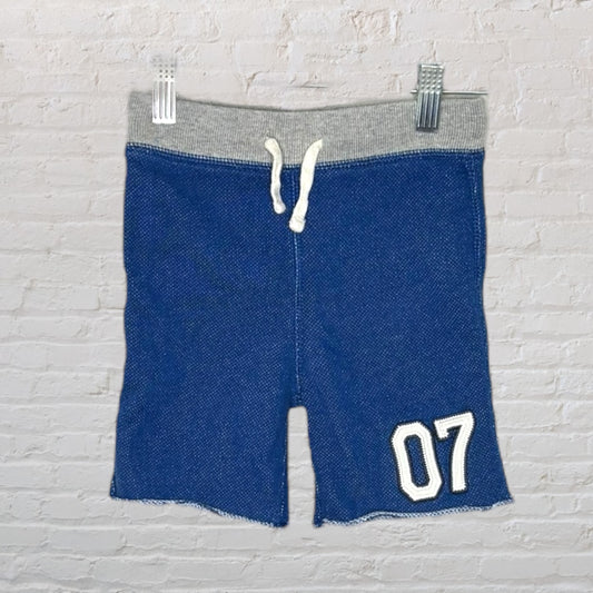 Blue knit shorts with grey waistband, drawstring, and '07' patch, hanging against a brick background.