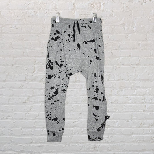 Grey sweatpants with black paint splatter design, featuring an elastic waistband and drawstring, hanging against a white brick wall.