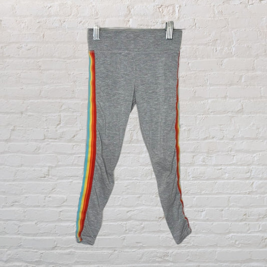 Gray leggings with colorful rainbow stripes down the sides, displayed on a hanger against a white brick wall.