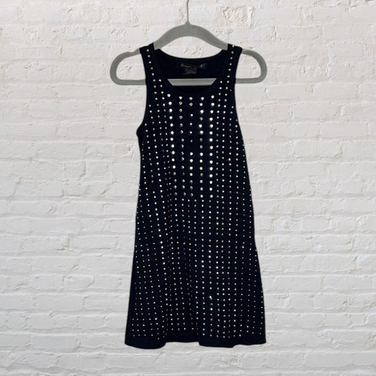 Black sleeveless dress with silver studs, hanging on a hanger against a white brick wall.
