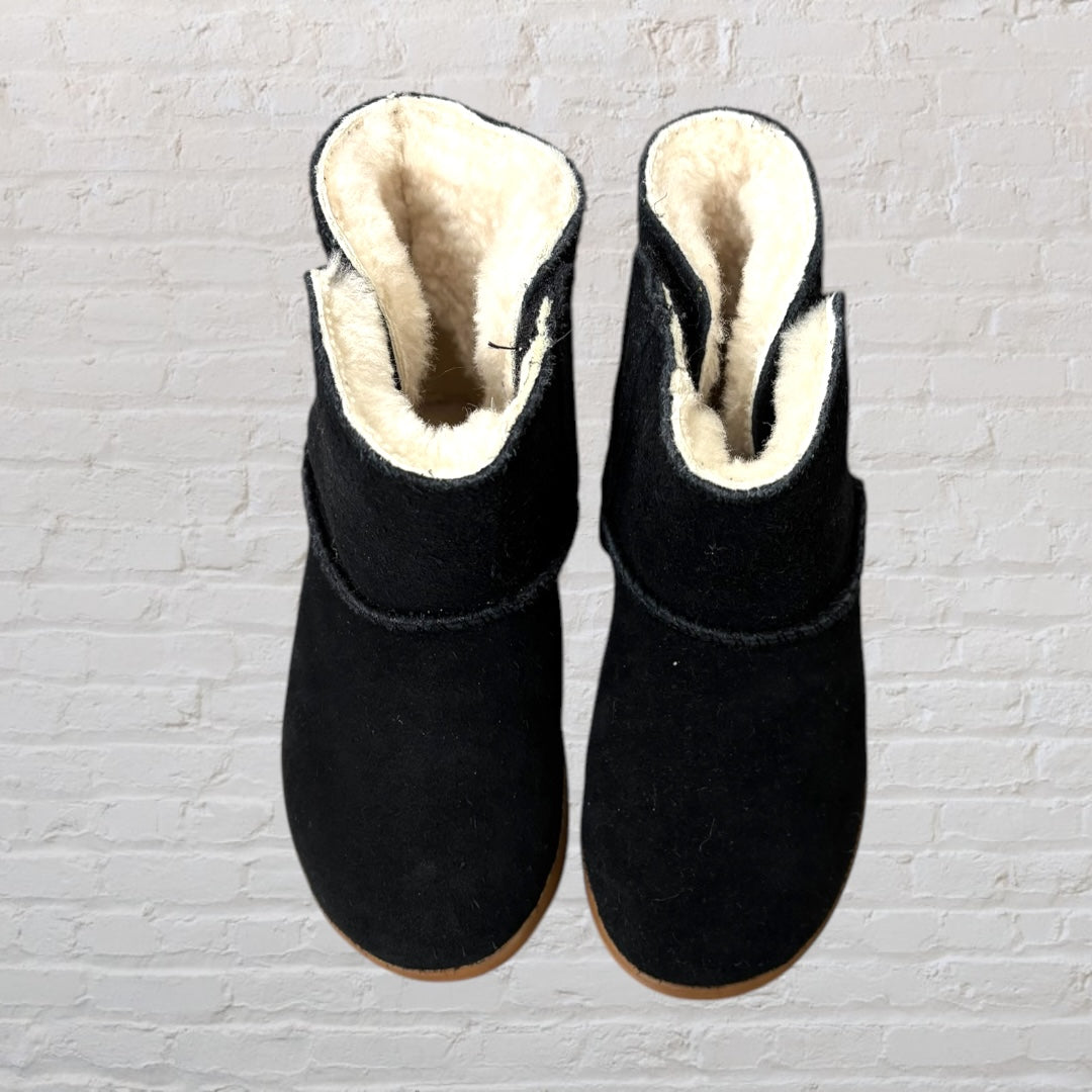 Black UGG boots with shearling lining and brown sole, side view against a brick wall background.