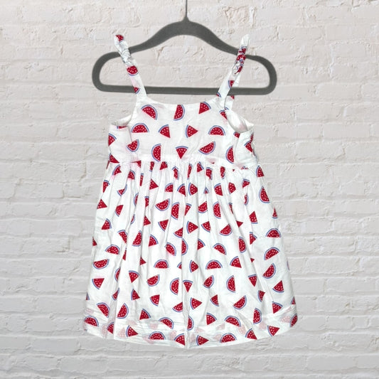 White sundress with watermelon print and adjustable straps displayed on a hanger against a brick wall.