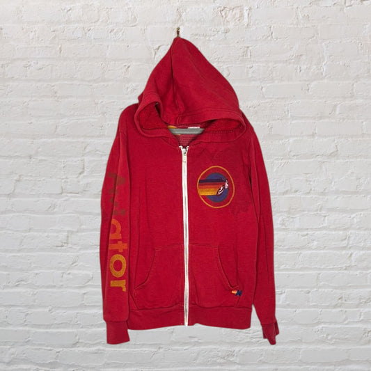 Red zip-up hoodie with a graphic patch on the chest and printed text on the sleeve, displayed against a white brick wall.