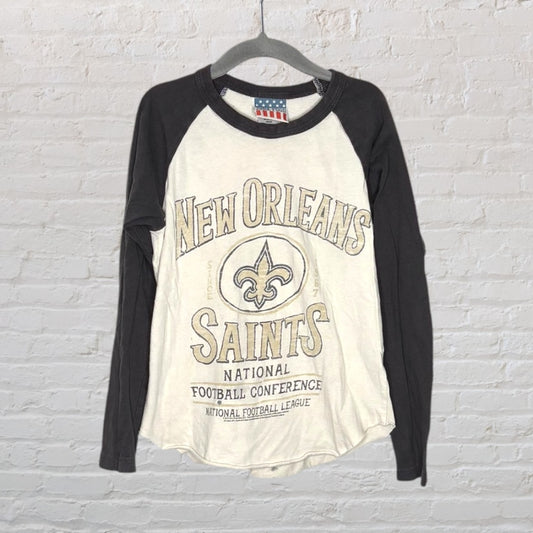 Cream and black long sleeve raglan T-shirt featuring New Orleans Saints graphics on the front, hanging against a brick wall.
