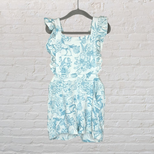 White and blue floral print romper with ruffle straps hanging on a hanger against a brick wall.
