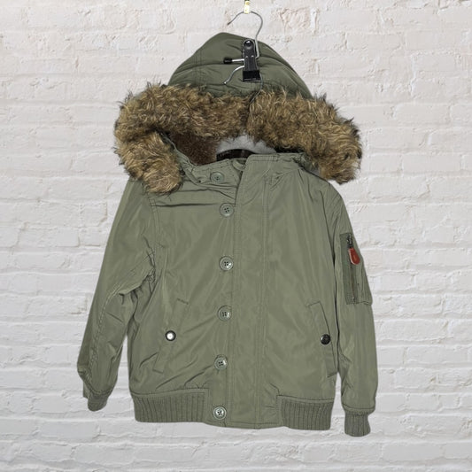 Olive green parka jacket with a faux fur hood, button closures, and ribbed cuffs, hanging on a hanger against a white brick wall.
