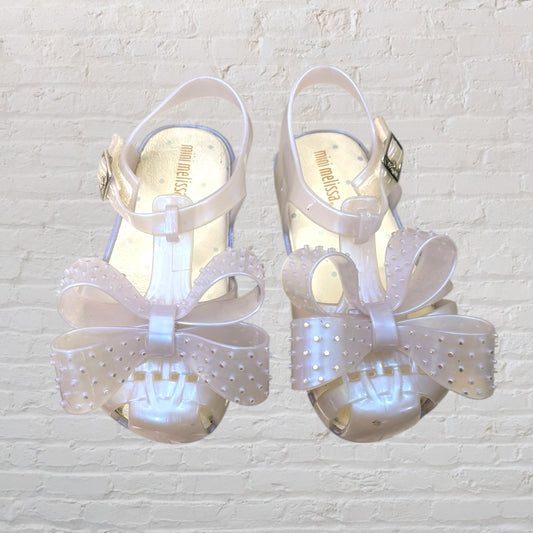 Pearl white Mini Melissa jelly sandals with large bow accents on a brick background.