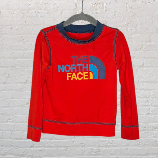 Red long sleeve tee by The North Face with a blue and yellow logo, hanging on a black hanger.