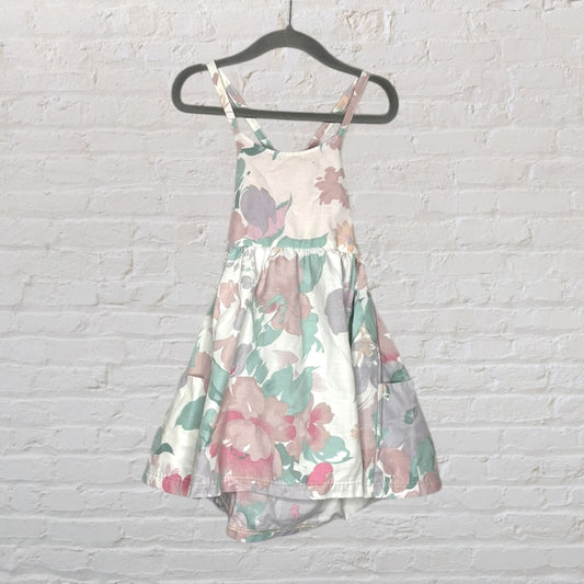 A sleeveless sundress with a floral print in pastel colors, hanging on a hanger against a white brick wall.