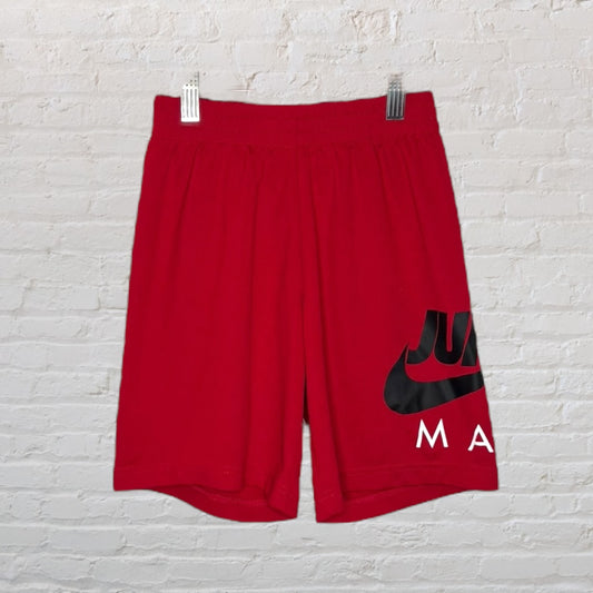 Red sports shorts with a large black and red graphic logo on the front, hanging against a white brick wall.