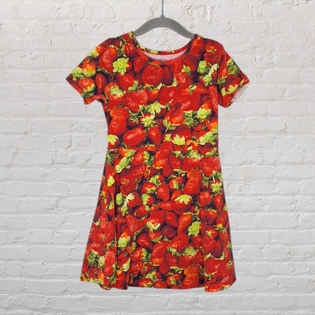 A short-sleeve dress featuring an all-over strawberry print with red and green colors, hanging against a white brick wall.