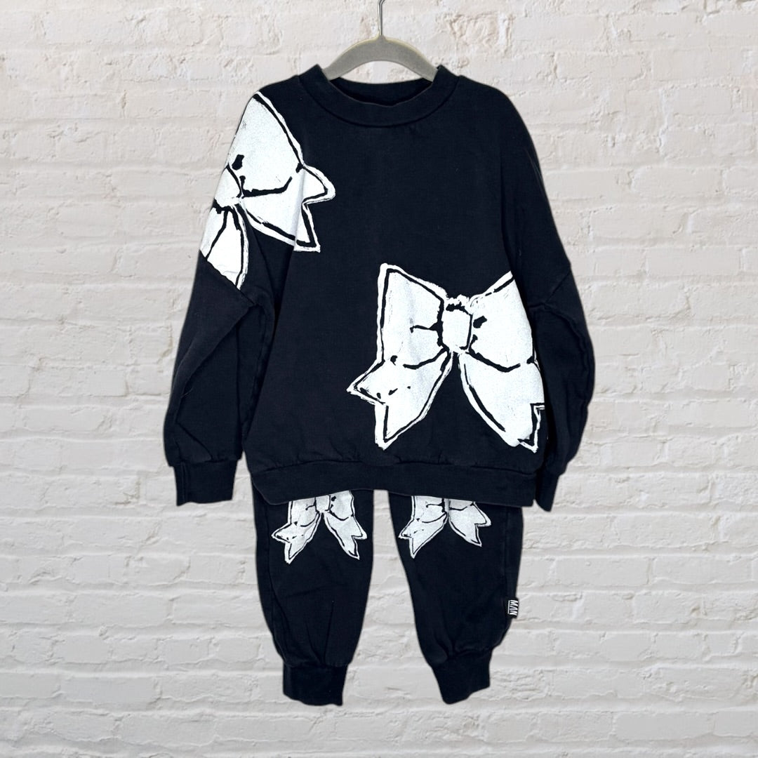 Black sweatshirt and pants set featuring large white bow graphics, displayed on a hanger against a brick wall.
