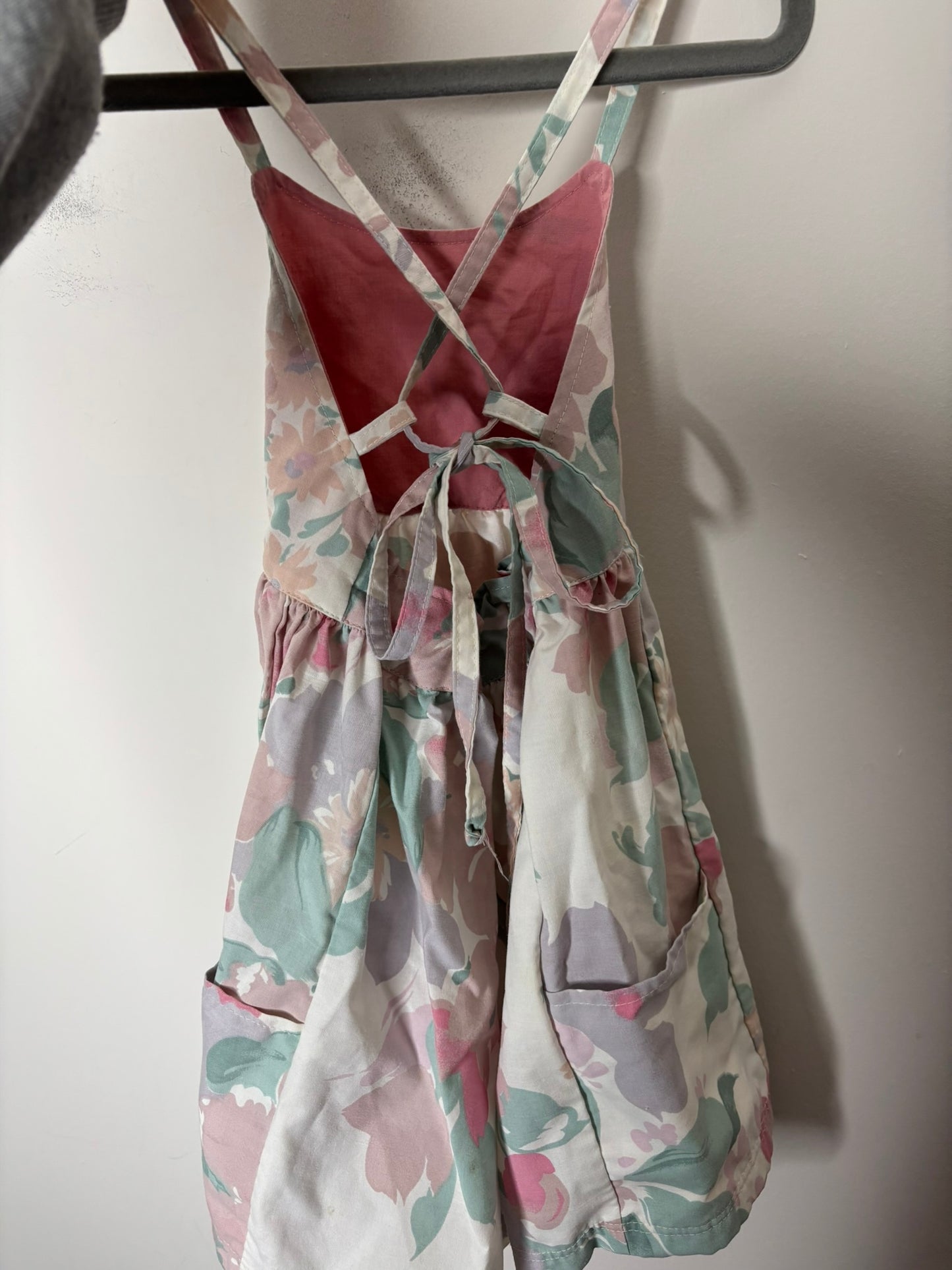 A sleeveless sundress with a floral print in pastel colors, hanging on a hanger against a white brick wall.