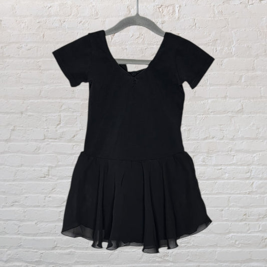 Black ballet leotard with short sleeves and attached skirt, hanging against a white brick wall.