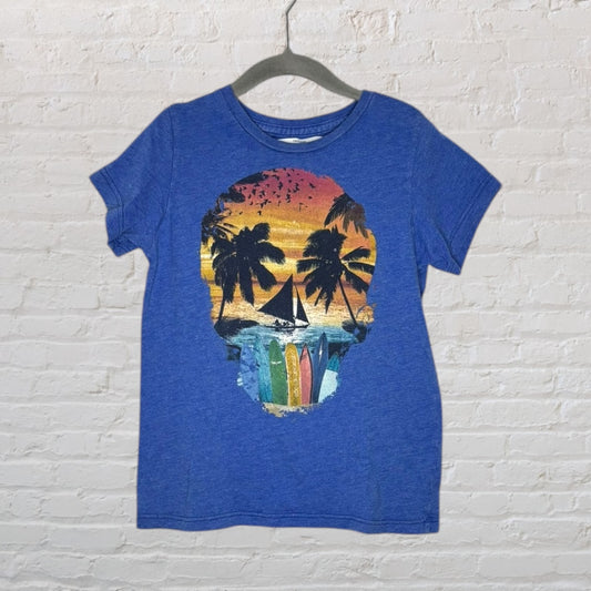 Blue T-shirt featuring a tropical skull graphic with a sunset, palm trees, and surfboards, hanging on a hanger against a brick wall.