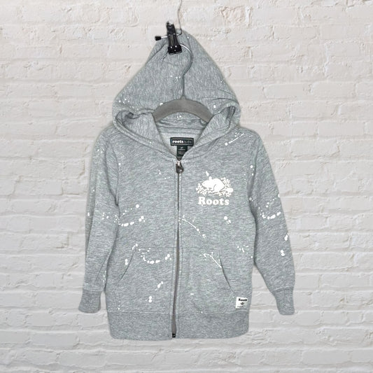 Grey Roots Kids zip-up hoodie with white splatter design and logo on chest, displayed against a brick wall.