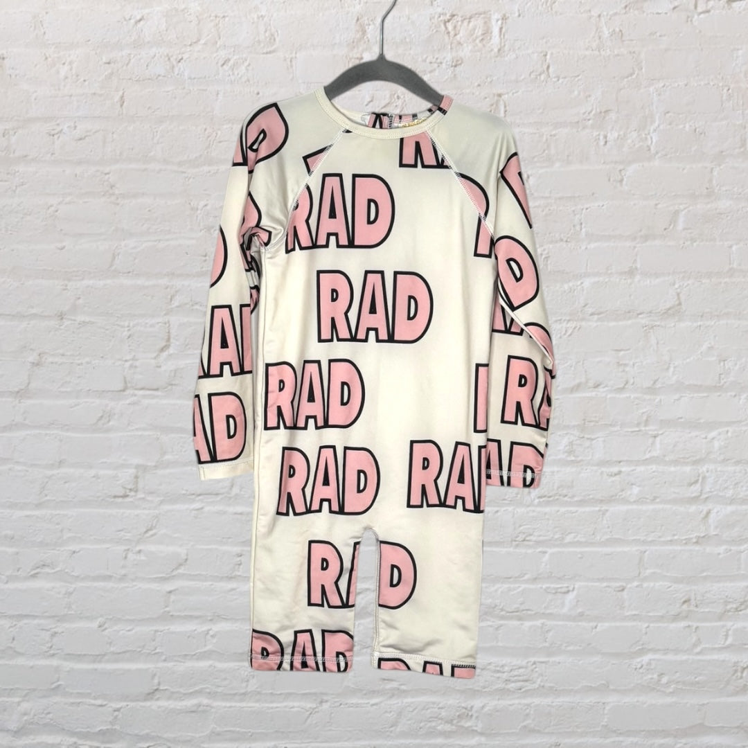 Long sleeve white romper with 'RAD' printed in pink all over, hanging against a white brick wall.