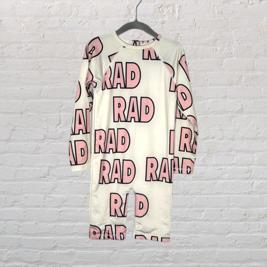 Long sleeve white romper with 'RAD' printed in pink all over, hanging against a white brick wall.