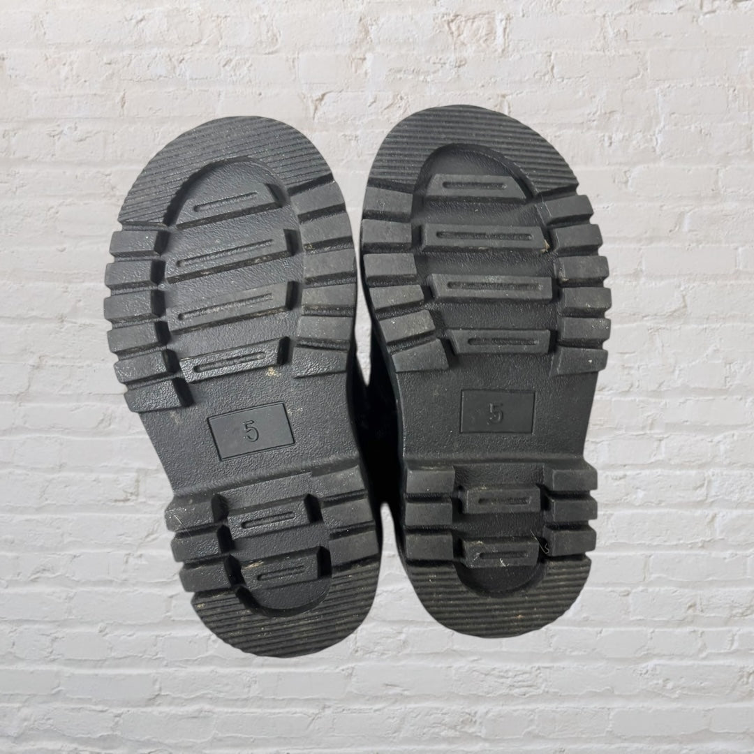 Pair of chunky black boots with silver buckle accents and a thick sole against a white brick background.