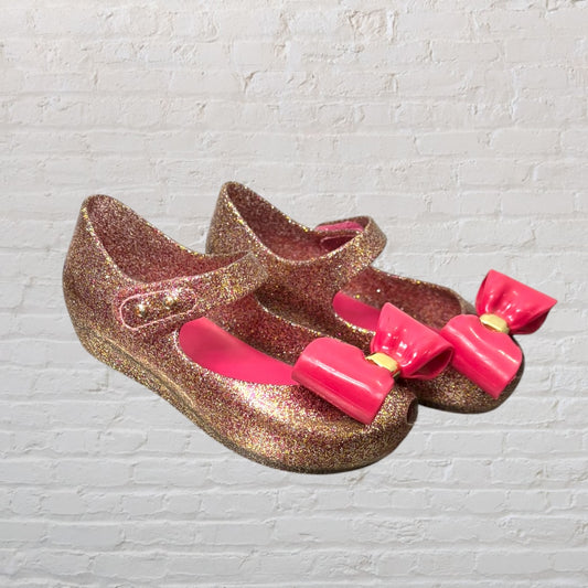 Pair of glittery gold Mary Jane shoes with pink bows on the toes, displayed against a white brick wall.