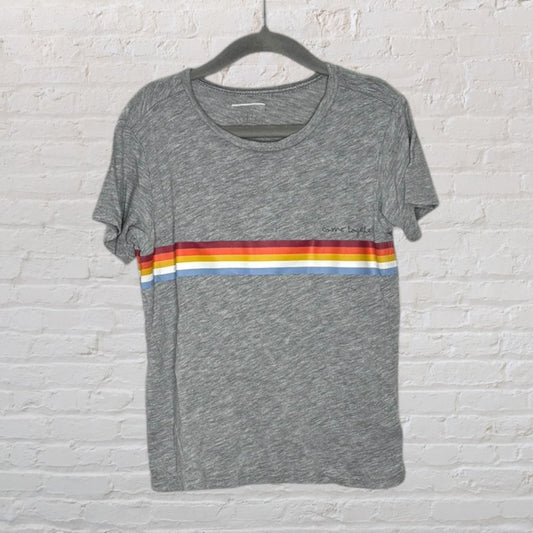 Grey T-shirt with colorful stripes and small print, hanging against a white brick background.