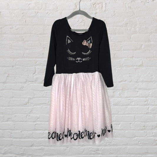 Black long sleeve dress with cat face design and pink tulle skirt, hanging on a hanger against a white brick wall.
